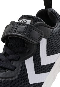 Schwarzer Sportschuh mit Mesh-Gewebe, weißem Zickzack-Seitendesign, schwarzen Schnürsenkeln und einem Leder-Klettverschluss am oberen Teil.