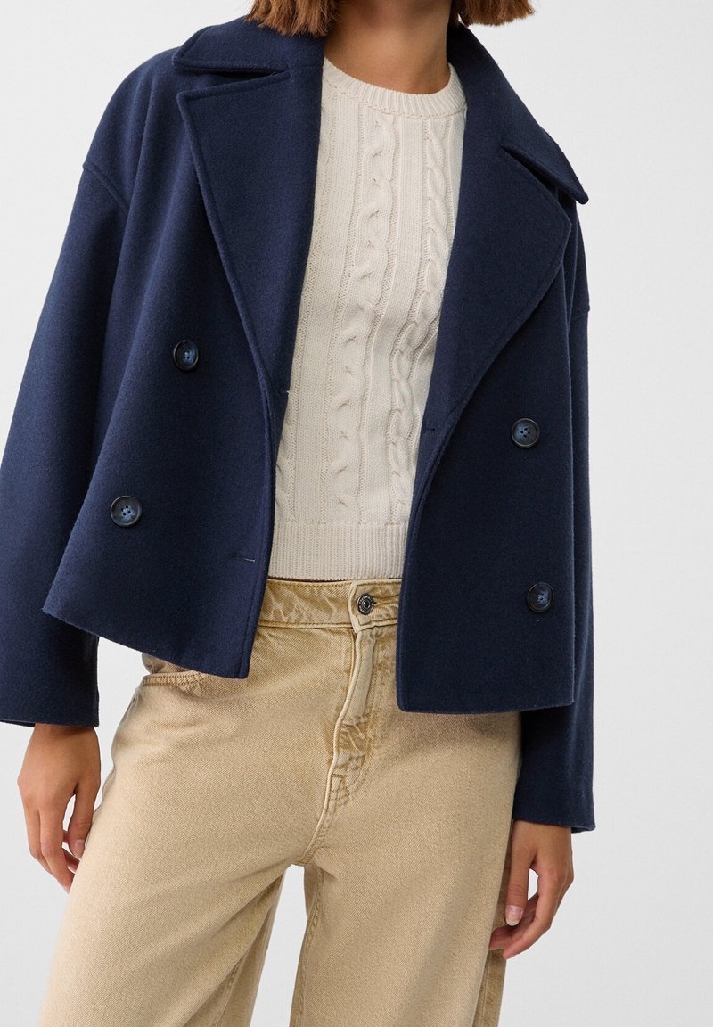 Marinblå kort kappa i ullblandning med slagkrage, dubbelknapprad och fickor med klaff, tillsammans med en krämfärgad kabelstickad tröja och beiga byxor.