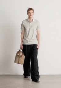 Ανοιχτό γκρι πόλο μπλουζάκι με κοντά μανίκια, μαύροι φαρδιοί παντελόνια και μια καφέ τσάντα τύπου duffel με καφέ λεπτομέρειες, σε φόντο απλό.