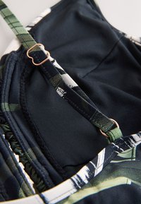 Bretelle fine réglable avec curseur doré sur un vêtement en tissu foncé à motif, montrant les détails de couture et la texture du tissu.