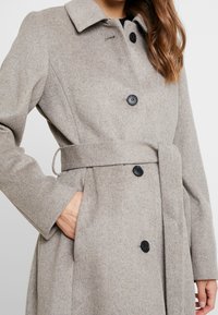 Abrigo de lana gris con un collar clásico, dos botones negros, bolsillos laterales y cinturón de tela a juego, con una textura suave y un diseño ajustado.