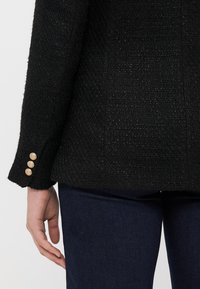 Juodas tekstūruotas švarkas su trimis auksiniais mygtukais ant rankovės, lygia medžiaga ir struktūruota siluetu. Dėvimas su tamsiomis džinsinėmis kelnėmis.