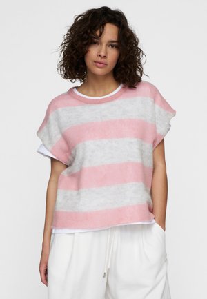 Donna con capelli ricci e scuri che indossa un maglione a maniche corte a righe rosa e grigie sopra una maglietta bianca, in posa con una mano nella tasca dei pantaloni bianchi.