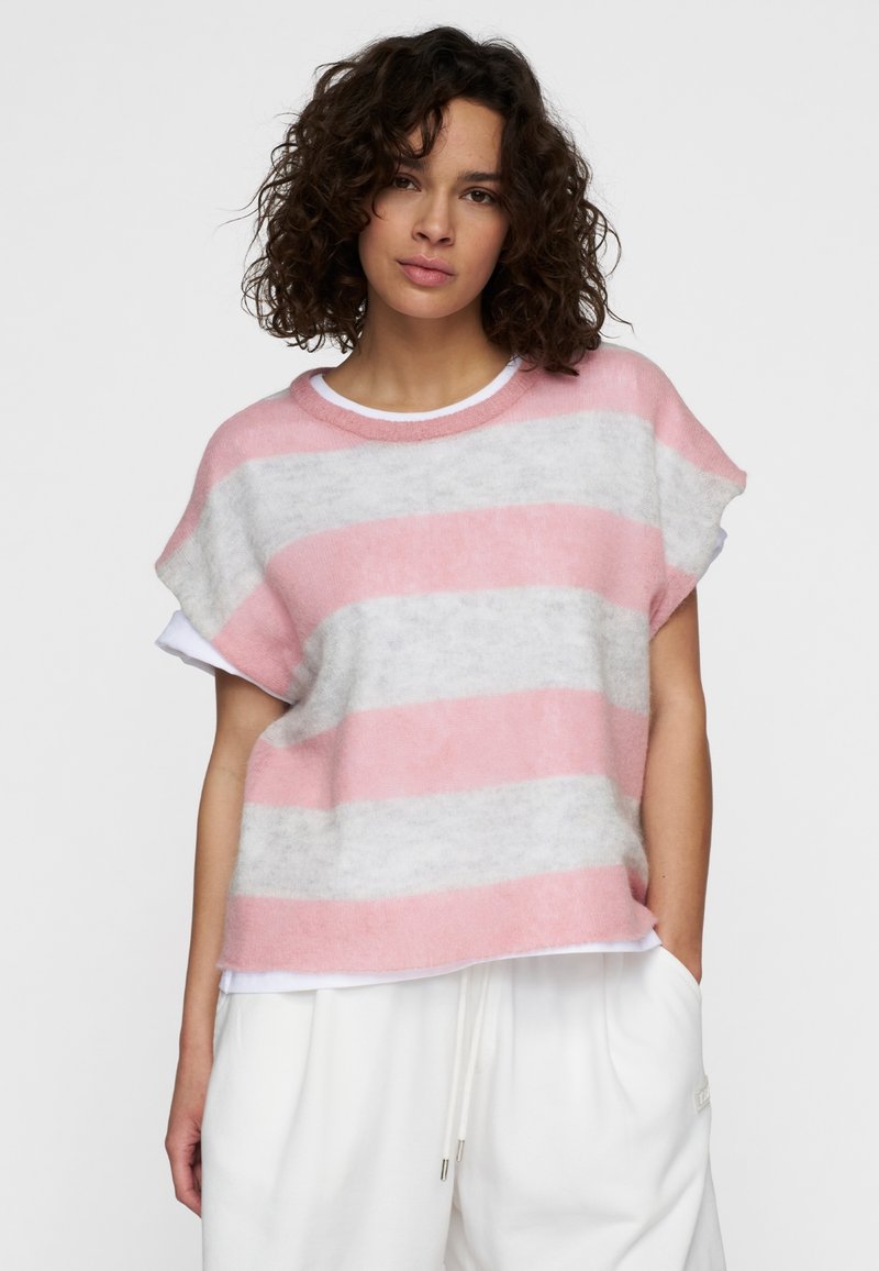 Mujer con cabello rizado y oscuro, vestida con un suéter de rayas rosa y gris de manga corta sobre una camisa blanca, posando con una mano en el bolsillo de unos pantalones blancos.