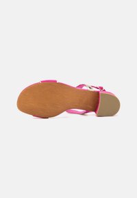 Pink sandal with a wooden sole, metallic buckle, and crisscross strap design. The sole features a textured grip for traction.