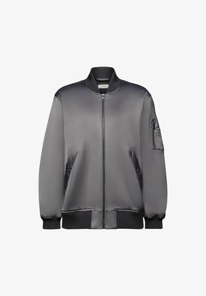 Chaqueta bomber gris sobre una sudadera con capucha naranja; minifalda negra con lentejuelas; botas de combate negras con cordones; bolso bandolera negro. Fondo verde.