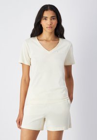 T-shirt a V con scollo leggero, di colore crema, maniche corte, in tessuto morbido e con branding minimale. Abbinato a pantaloni corti crema coordinati.