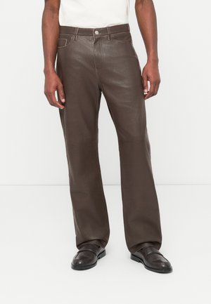 Muž nosiaci tmavohnedé kožené nohavice a zladené tmavohnedé kožené topánky, stojaci na bielom pozadí.