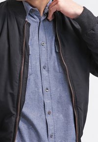 Black bomber jacket with ribbed cuffs, worn over a blue patterned button-up shirt featuring a subtle floral design and chest pocket.