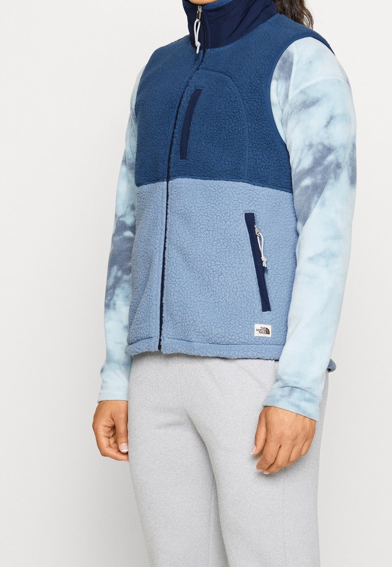 Osoba v dvoubarevném modrém fleecovém vestě přes světle modré tričko s dlouhým rukávem v batikovém stylu a světle šedých kalhotách, stojící s uvolněnými pažemi.