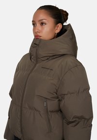 Giacca a vento marrone con colletto alto, texture trapuntata orizzontale, zip frontale e tasche laterali. Finitura del tessuto liscia con dettaglio del logo.