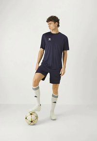 Jūrinės sporto marškinėliai su atitinkamomis šortais, pagaminti iš lengvos medžiagos. Baltos kojinės su juodomis juostelėmis. Žaidėjas stovi su spalvingu futbolo kamuoliu.