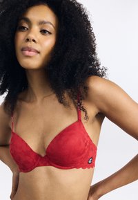 Punainen pitsinen bralette, jossa on kukkakuosi, tukeva alustuet ja säädettävät hihnat. Hapsureunat ja pieni logotieto vyössä.