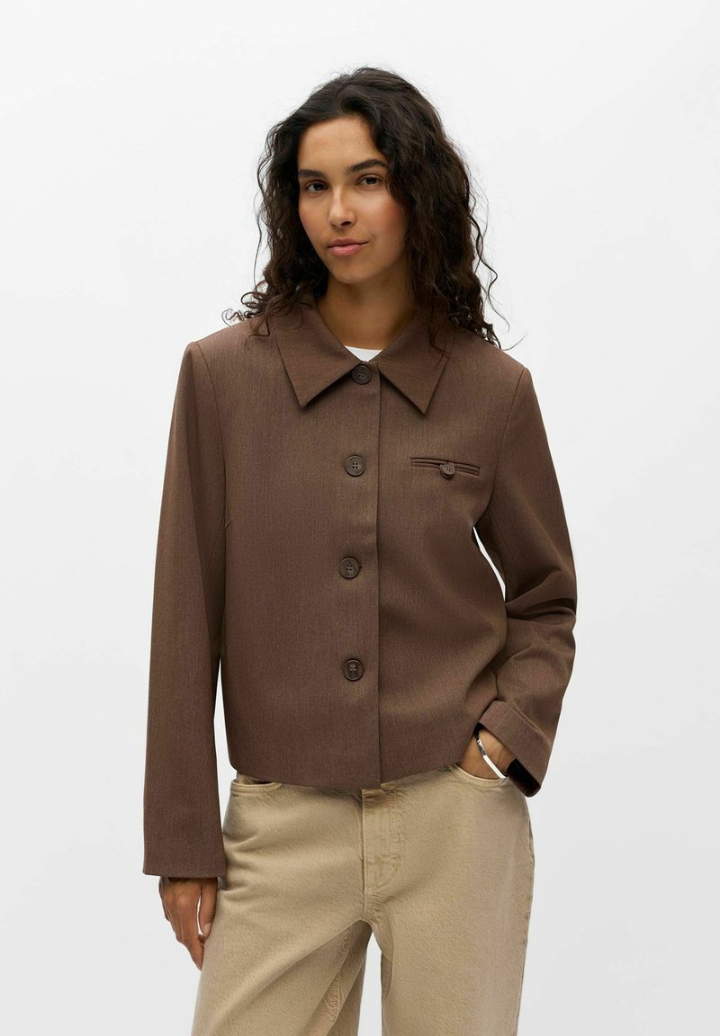 Femme aux cheveux bouclés et foncés portant une veste marron boutonnée et un pantalon beige, debout avec une main dans la poche, sur fond blanc.
