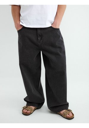 Person trägt lose schwarze Jeans mit Händen in den Taschen und beigen Sandalen mit Schnalle auf schlichtem hellem Hintergrund.