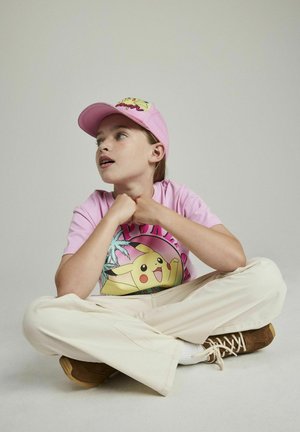 Casquette rose avec un logo, T-shirt rose avec un graphique de Pikachu, et pantalon large beige. Des bottes marron complètent la tenue.