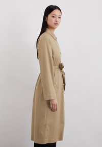 Trench coat beige a maniche lunghe con cintura, colletto, tasche laterali e una vestibilità rilassata, realizzato in un tessuto liscio.