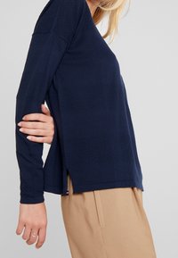 Granatowy top z długim rękawem z teksturowanymi poziomymi paskami, o luźnym kroju i rozcięciach po bokach. Wykonany z miękkiego materiału, odpowiedni do codziennego noszenia.