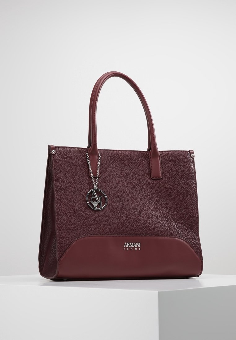 Bordowa skórzana torba o fakturowanej powierzchni, z dwoma uchwytami i srebrnym zawieszką z logo. Posiada gładkie dno z tłoczonym nazwiskiem marki.