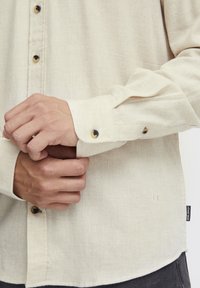 Chemise à manches longues beige clair en tissu texturé, ornée de boutons noirs et d'une petite étiquette logo sur le côté près de l'ourlet.
