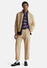 Chaqueta beige, camisa azul marino, camiseta a rayas marrón debajo, pantalones beige, mocasines negros, calcetines grises. Conjunto casual en capas con texturas contrastantes.