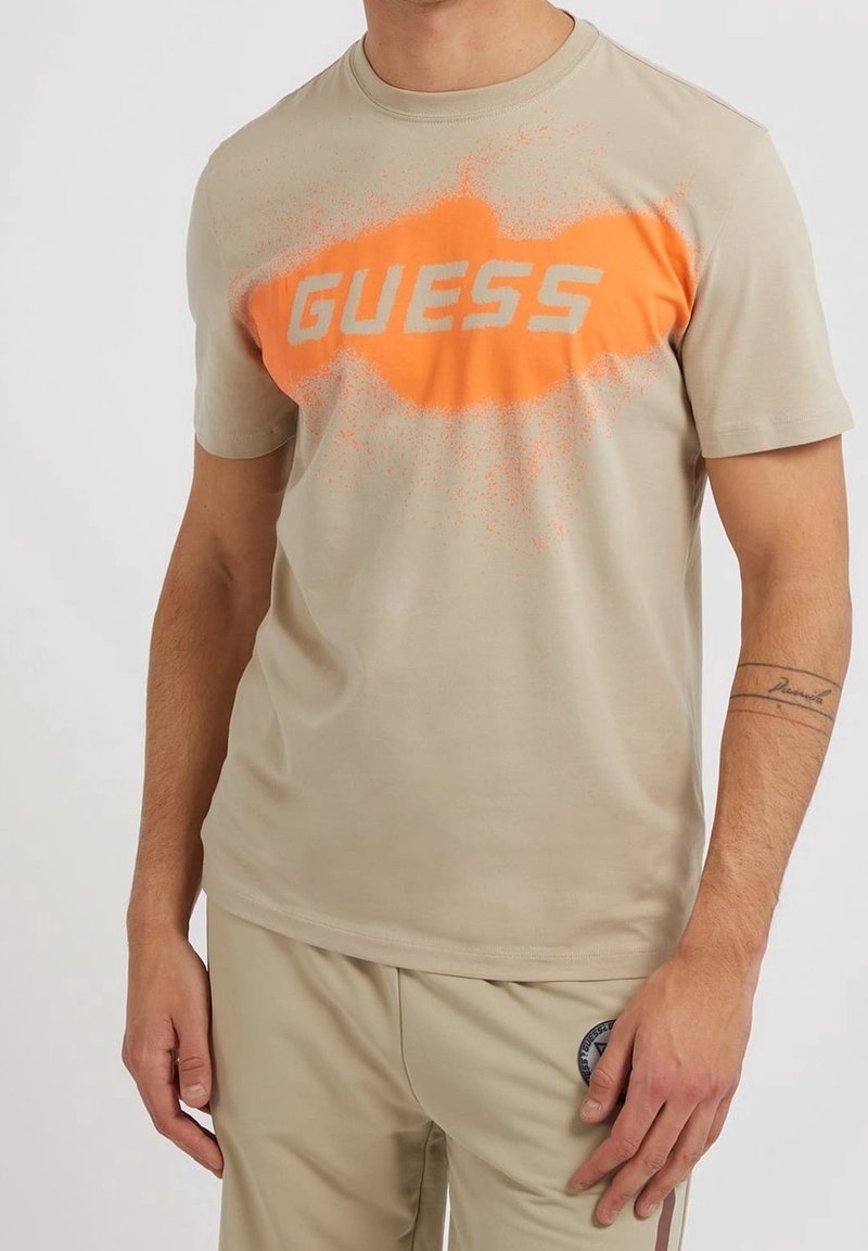 T-shirt en coton beige avec un col rond. Présente un grand logo "GUESS" orange avec un effet texturé, de type pulvérisé, au centre.