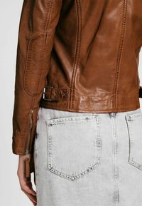 Giacca di pelle marrone con una vestibilità strutturata, superficie testurizzata, zip laterale e dettaglio con cintura in vita, abbinata a una gonna di jeans chiara.