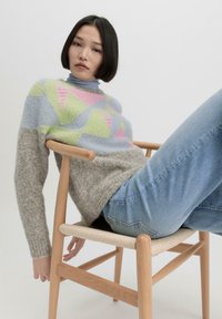 Gebreid sweater met geometrische patronen in groen, roze en blauw, gecombineerd met lichtblauwe jeans. Model zit op een houten stoel.