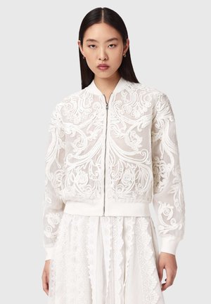 Junge Frau mit langen dunklen Haaren, die eine weiße, durchsichtige Bomberjacke mit kunstvoller Stickerei und einen passenden Spitzenrock trägt.