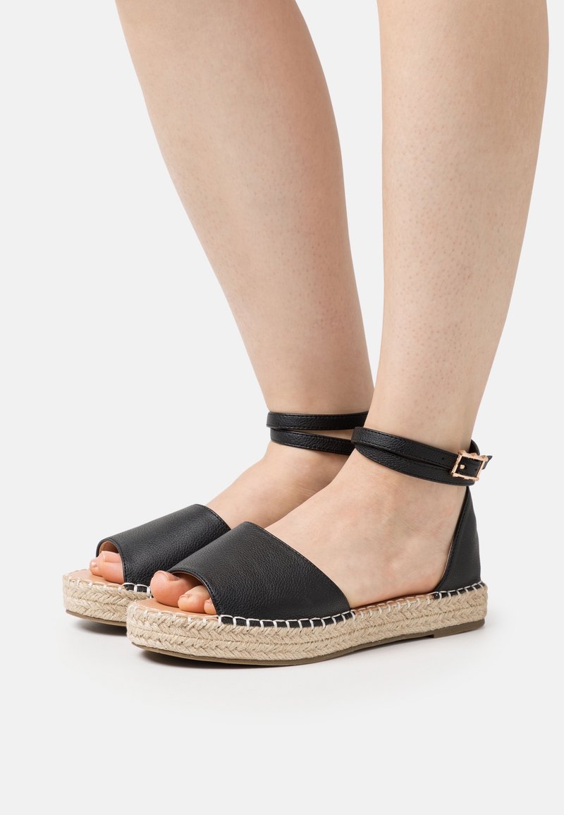 Pieds portant des sandales espadrilles en cuir noir avec brides à la cheville et semelles plateformes en jute tressée sur un fond clair.