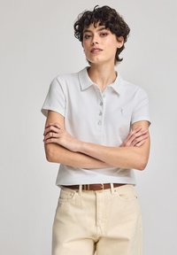 Vit polo skjorta med korta ärmar, krage och ljusblå knappar. Bärs med ljusbeiga jeans och ett brunt bälte.