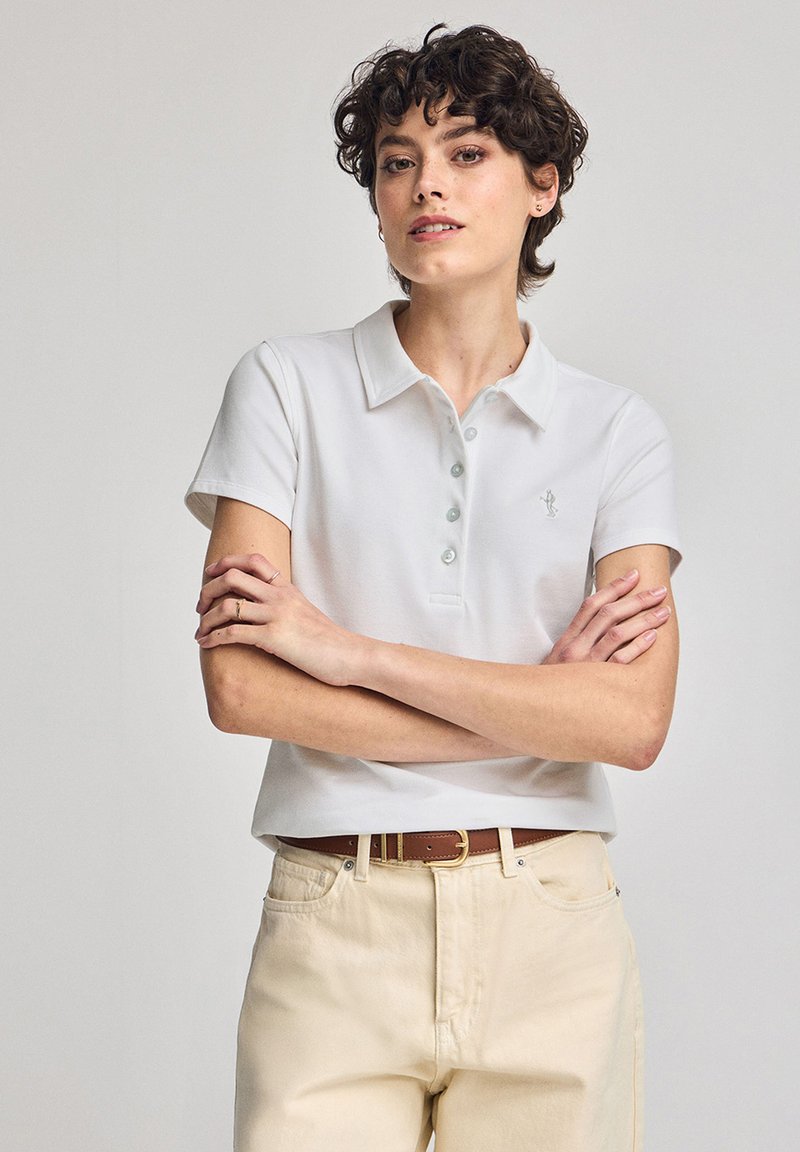 Vit polo skjorta med korta ärmar, krage och ljusblå knappar. Bärs med ljusbeiga jeans och ett brunt bälte.