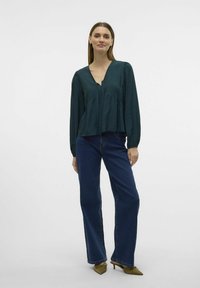 Teal bluse med V-udskæring og plisserede detaljer, parret med højhalsede mørkeblå jeans og olivengrønne sko. Glat stof, afslappet pasform.