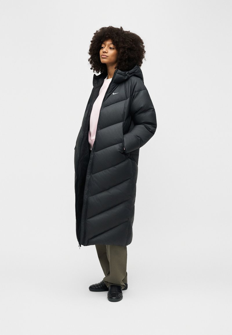 Frau mit lockigem Haar, die einen langen schwarzen Nike-Steppmantel über einem hellrosa Oberteil, olivgrünen Hosen und schwarzen Turnschuhen trägt, steht mit den Händen in den Taschen.