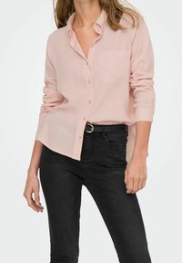 Camisa de botones de color rosa claro con un bolsillo en el pecho, mangas largas y un corte relajado, combinada con unos pantalones vaqueros ajustados oscuros. Textura de tela suave.