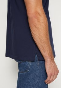 Marineblaue T-Shirt mit kurzen Ärmeln, aus weichem Baumwollstoff. Der Saum hat seitliche Schlitze für mehr Bewegungsfreiheit und wird mit blauen Jeans kombiniert.