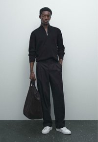 Zwarte ribgebreide sweater met kwart ritssluiting, ruimvallende zwarte broek, witte sneakers en een donkerbruine suède tote bag. Smooth texturen, minimale vormgeving.