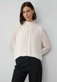 Woman wearing white high-neck blouse with long sleeves and black pants, standing with hands behind her back against a plain light wall.