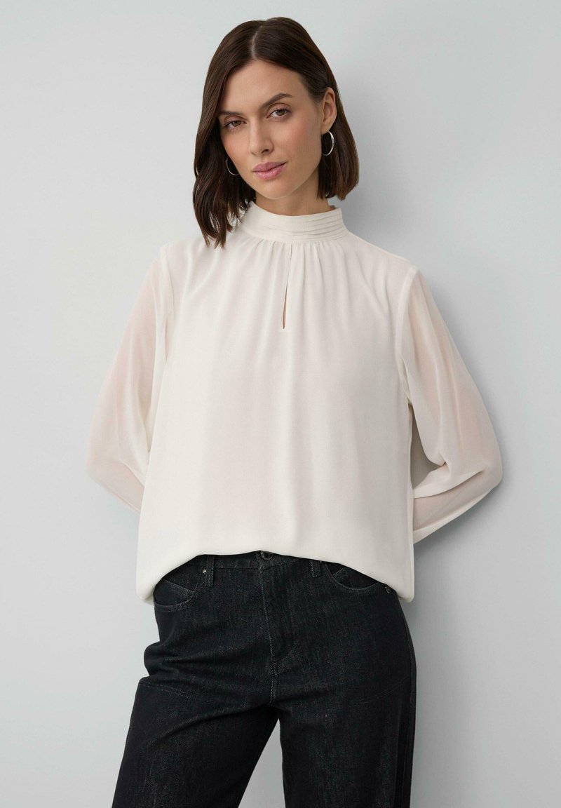 Woman wearing white high-neck blouse with long sleeves and black pants, standing with hands behind her back against a plain light wall.