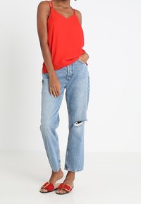 Haut rouge à fines bretelles, jean bleu clair taille haute déchiré, et sandales rouges à glissière avec des accents de boucle dorée. Design simple et coupe décontractée.