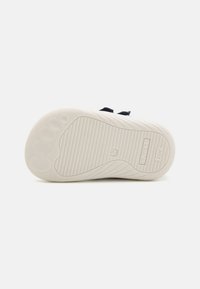 Weiße Gummisohle mit strukturiertem Profil, mit einem glatten, geschwungenen Design und geprägtem Logo. Dunkelblaue Textiloberseite mit Klettverschluss.