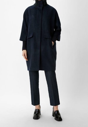 Femme portant un manteau bleu marine foncé à larges manches, un pantalon sombre et des mocassins noirs en cuir verni à talon bloc, debout sur fond blanc.