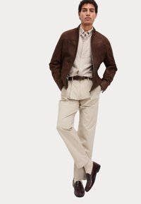 Brun mockajacka över en ljusbeige skjorta, kombinerat med beige byxor och mörkbruna loafers, som har ett enkelt bälte och en avslappnad passform.