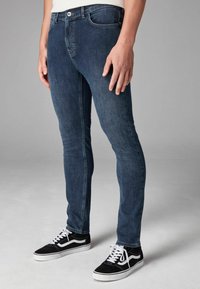 Sötét kék farmernadrág slim fit szabással, elülső zsebekkel, gombos cipzárral és letisztult megjelenéssel. Fekete sportcipővel párosítva.