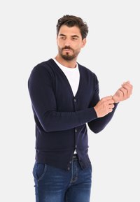 Cardigan blu navy realizzato in tessuto a maglia. Presenta una scollatura a V, maniche lunghe e chiusura con bottoni. Indossato con una maglietta bianca e jeans blu.