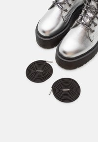 Botas de tobillo de cuero plateado con suelas gruesas negras y ojales plateados, acompañadas de dos cordones negros enrollados con puntas de metal.