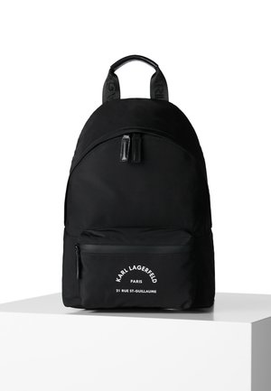 Mochila negra de Karl Lagerfeld con bolsillo frontal con cremallera y texto de logo blanco, de pie sobre un pedestal blanco contra un fondo blanco.