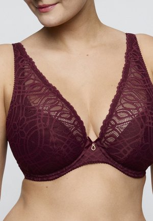 Vrouw die een diep bordeauxkleurige kanten beha draagt met ingewikkelde patronen en een kleine gouden druppelhanger aan de voorkant in het midden.