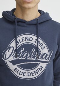 Kék kapucnis pulóver, fehér kör alakú grafikával, rajta az "Eredeti Kék Farmer" felirattal és kopottas kinézettel. Állítható zsinóros kapucni.