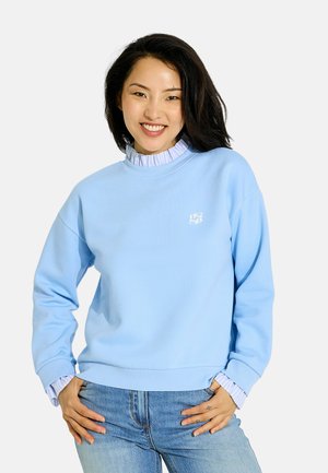 Mujer sonriente con cabello negro lleva una sudadera azul claro sobre una camisa blanca con volantes y vaqueros azul claro, con las manos apoyadas en las caderas.